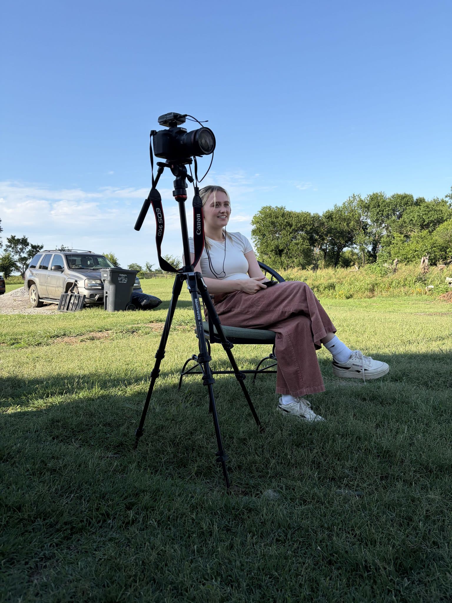 An interview subject sits for a portrait on a grassy field, tripod at the ready.
