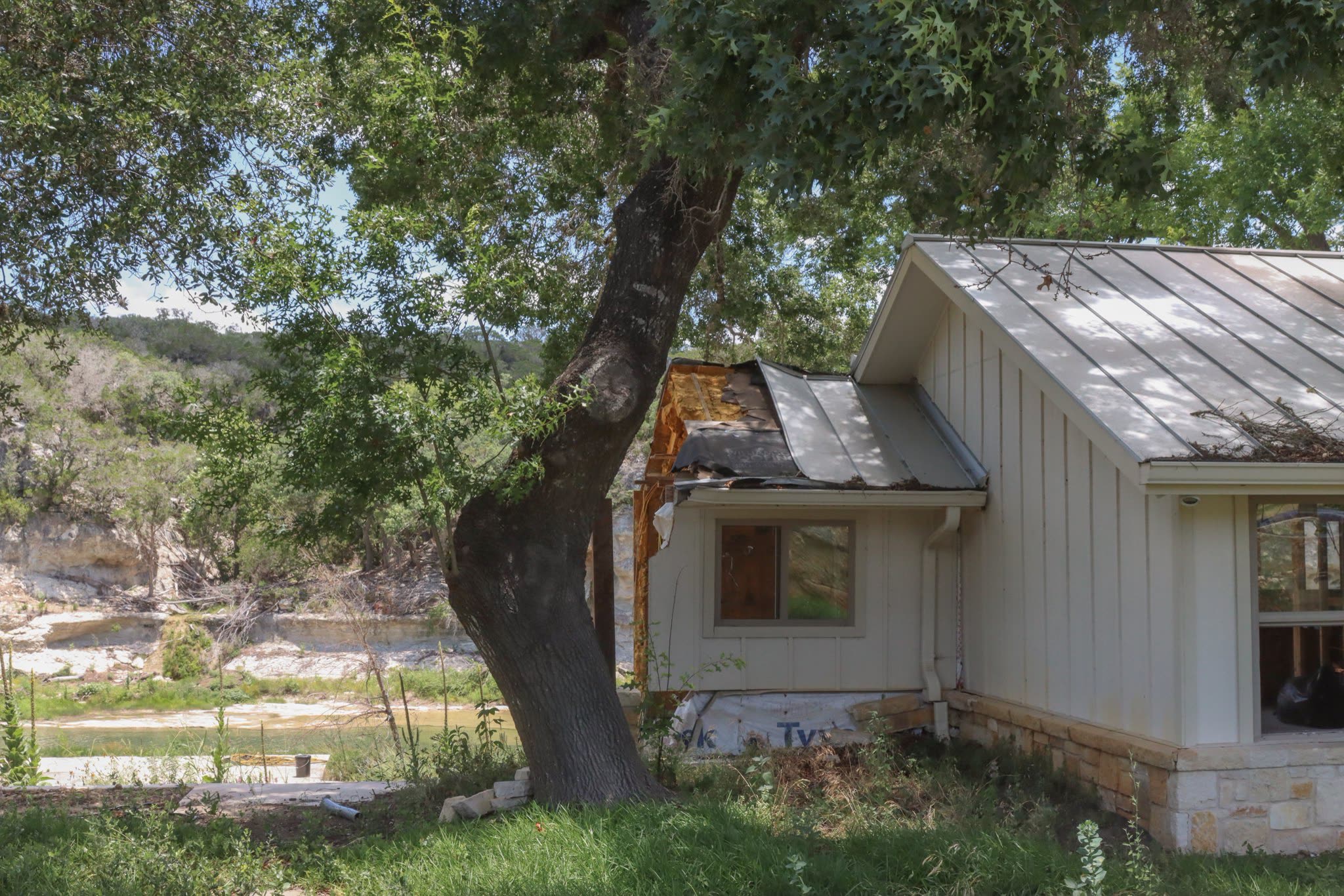 A damaged house sits split and sagging beneath a large tree near the river.